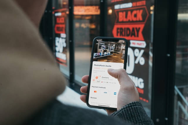Photo of a boy holding a phone with the location of a shop and a glass window covered in Black Friday stickers in the background. 